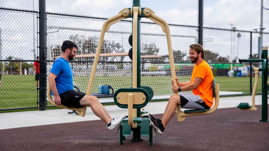 Two men exercising on a 4-Person Leg Press with a soccer field in the background