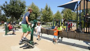 A man exercising on a Stepper talking to two children with a playground in the background.