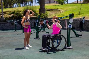 A woman in a wheelchair exercising on the Accessible Shoulder Press and another woman to her left talking to her at an outdoor gym.