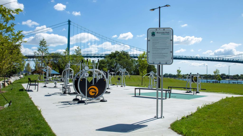 An outdoor gym with a sign and 2-Person Cross Country Ski in the foreground. A bridge and blue sky is the background.